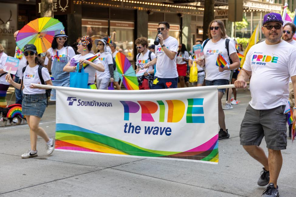 Sound Transit reps walk through the Pride parade holding a banner that reads, "Pride the Wave"