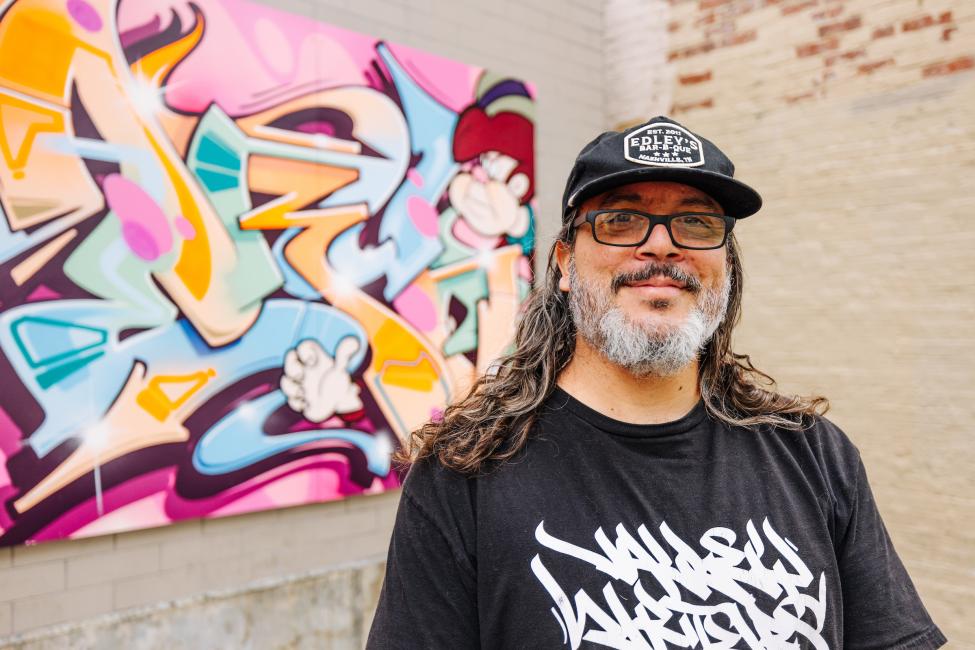 Danny Melbihess wears a black T-shirt and hat and smiles with a piece of colorful graffiti-style art in the background