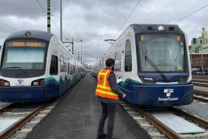 A person wearing an orange vest stands between two Link light rail vehicles