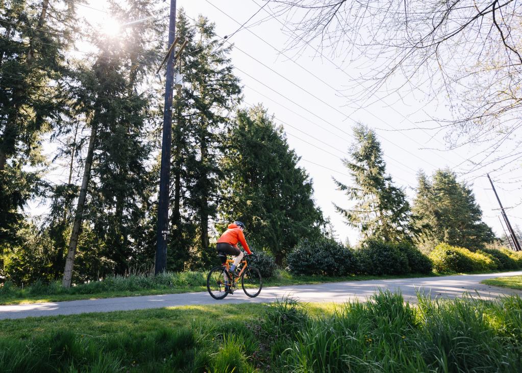 A cyclist rides on the Interurban Trail on a sunny day, with green trees in the background.